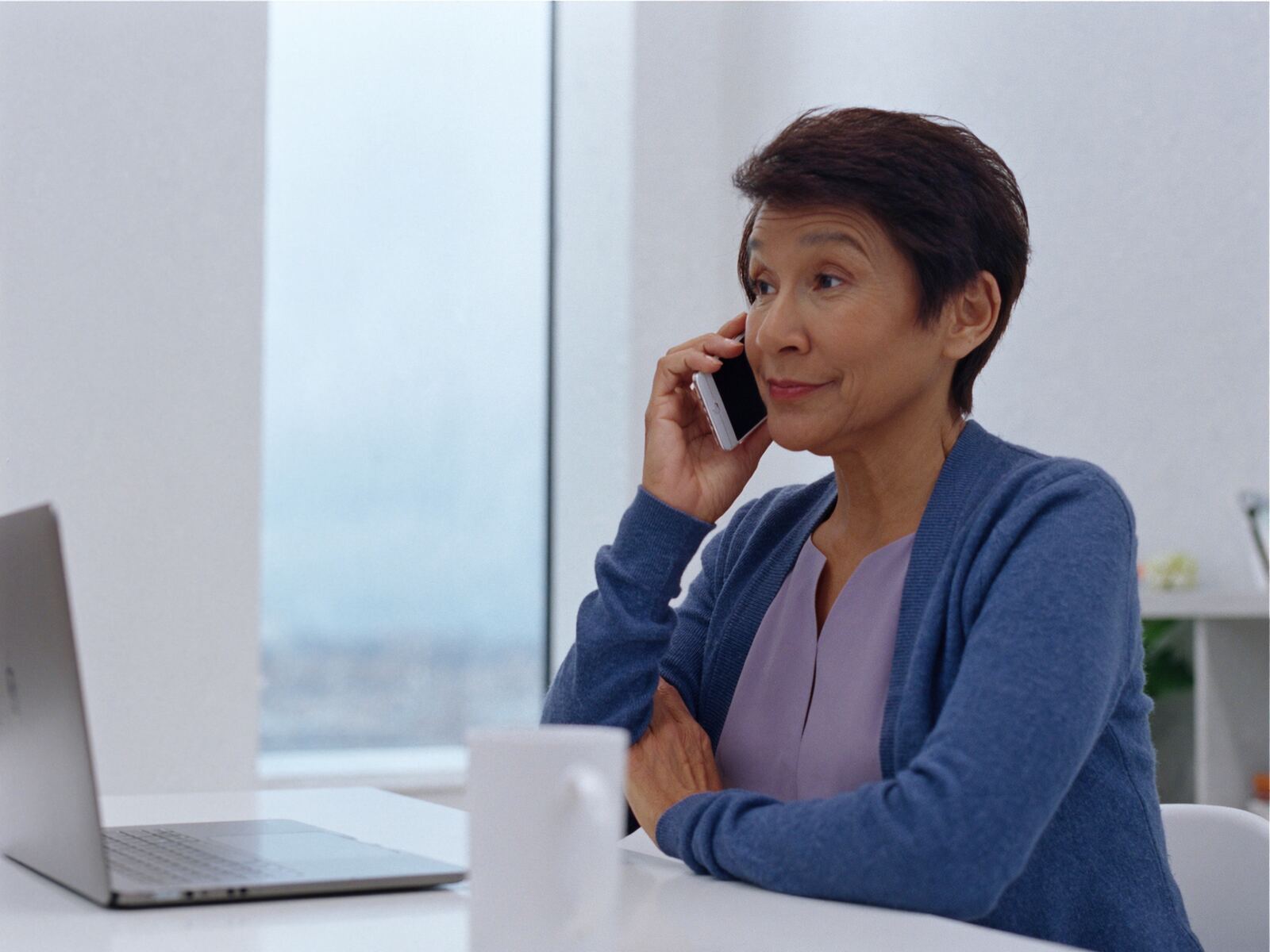 A ministry responder at a white desk beside a bright window, a slim phone to her ear, smiling as she listens, wearing a denim blue cardigan over a soft periwinkle top