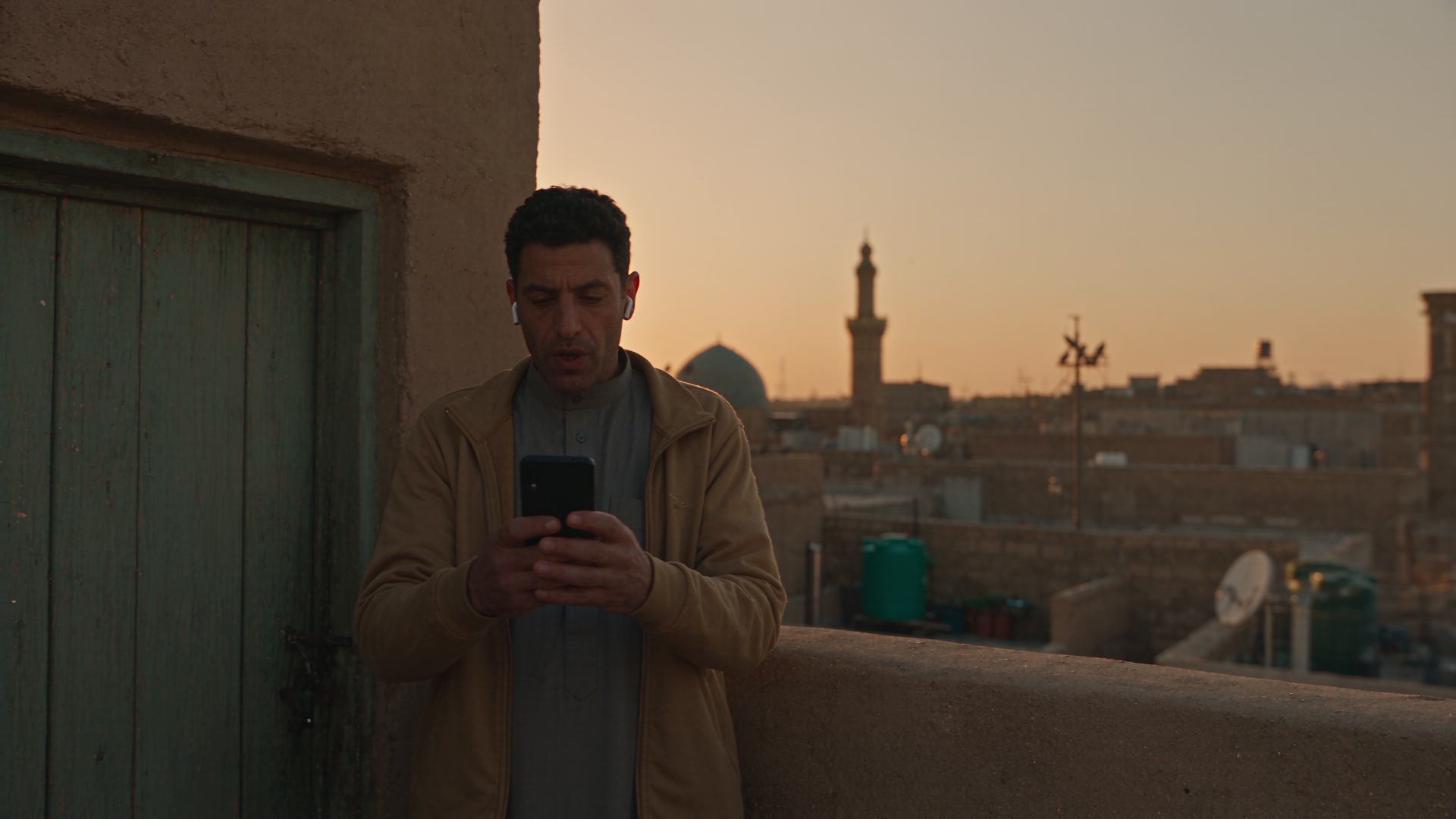 A man on a rooftop at dawn in a Middle Eastern city, on a quiet call, the city waking behind him