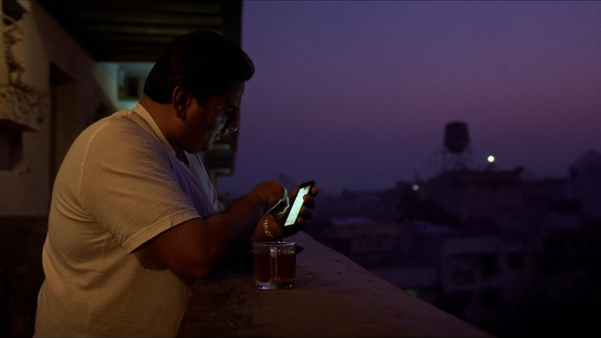 A man on an apartment balcony at dusk, reading a long reply on his phone, tea on the rail