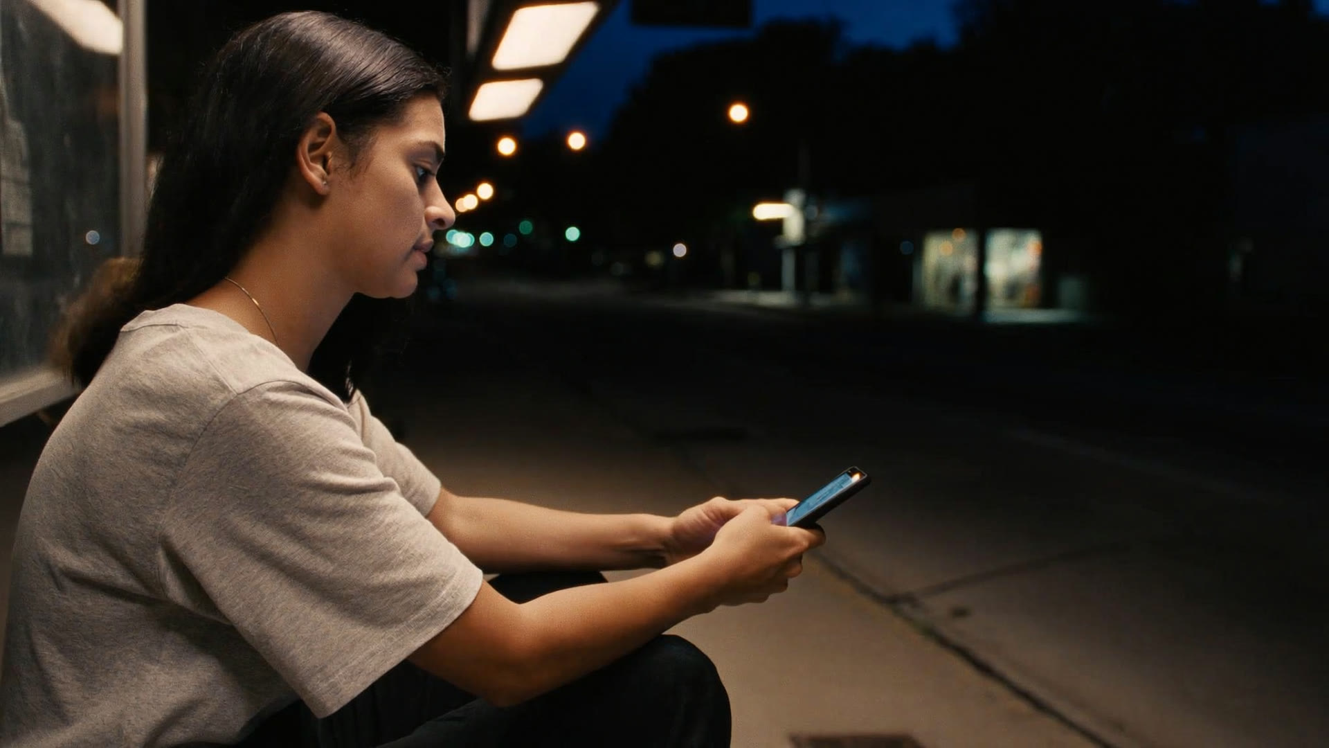 A woman sitting alone at a bus shelter at night, reading a reply on her phone