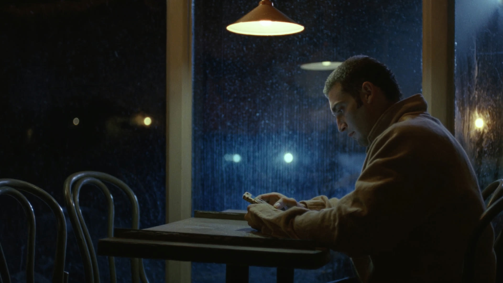 A man at a café table by a rainy window at night, reading his phone