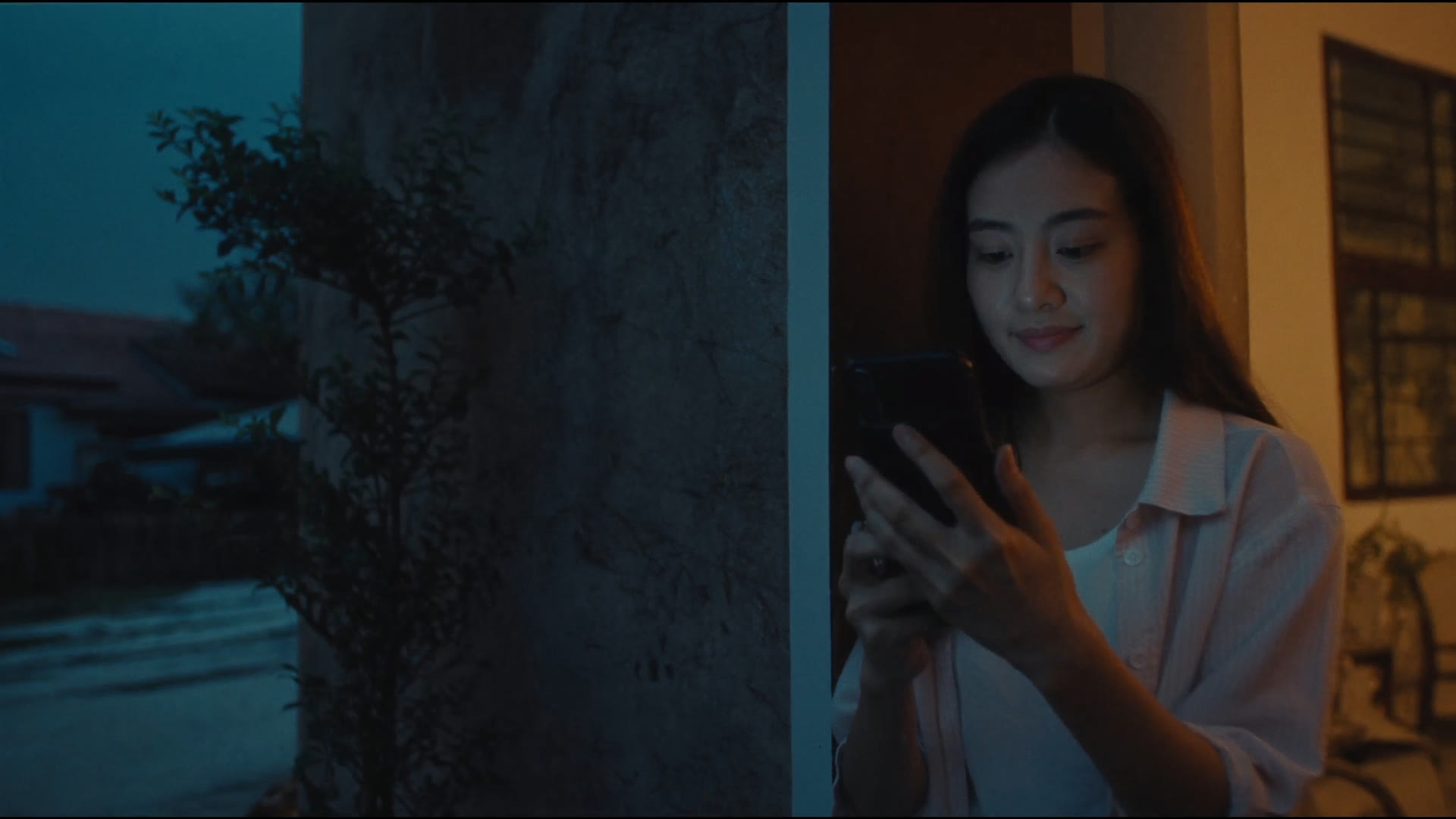 A woman in the lit doorway of her house at dusk, smiling at a reply on her phone