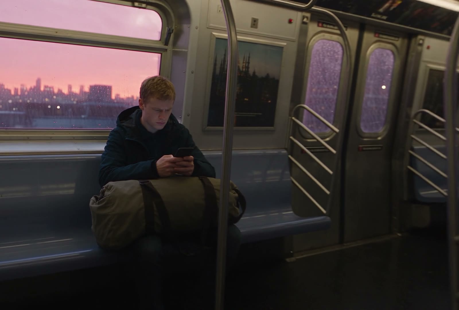 A young man on a train at dusk, reading his phone, a packed bag at his feet
